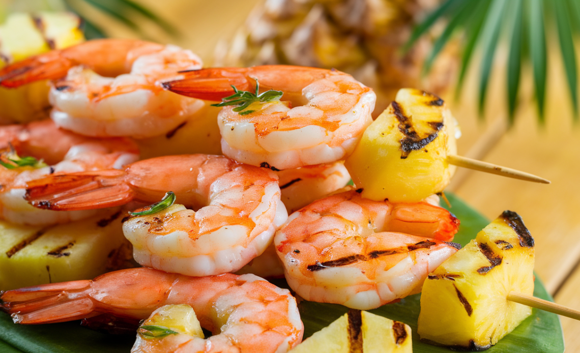 Grilled shrimp and pineapple pieces threaded on skewers, charred and glazed, served on a rustic wooden platter.